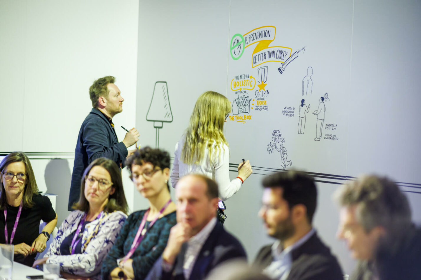 Graphic recorders illustrating ideas on a wall during a live workshop with participants watching the visual process unfold.