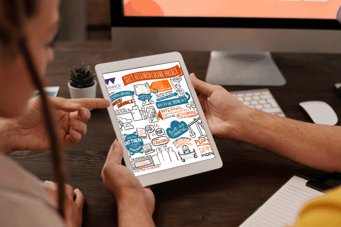 Hands holding a tablet showing a graphic recording from the SELCS Research Culture Project, demonstrating how event visuals can be reused digitally.